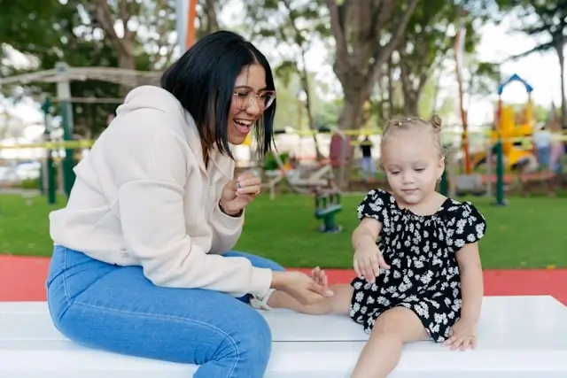 A smiling young woman and a little girl sitting together at a playground, playing and enjoying time outdoors.