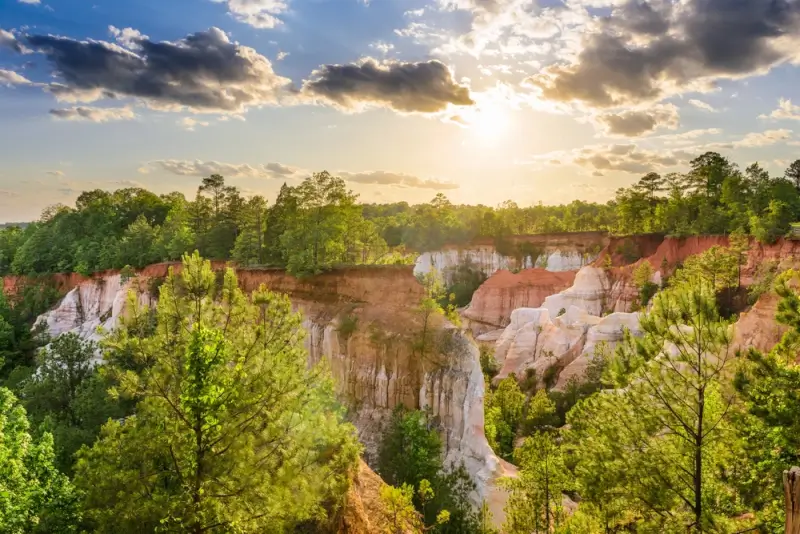 Providence Canyon near Columbus Georgia featuring dramatic red, orange and white rock formations with pine trees and sunlight breaking through clouds