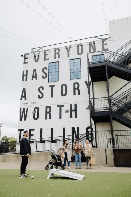 A group of adults with a stroller standing outside a large building with bold lettering that reads “Everyone Has a Story Worth Telling,” with outdoor steps and string lights above.
