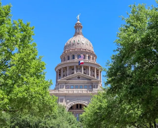The Texas State Capitol building framed by green trees on a bright sunny day with a clear blue sky.