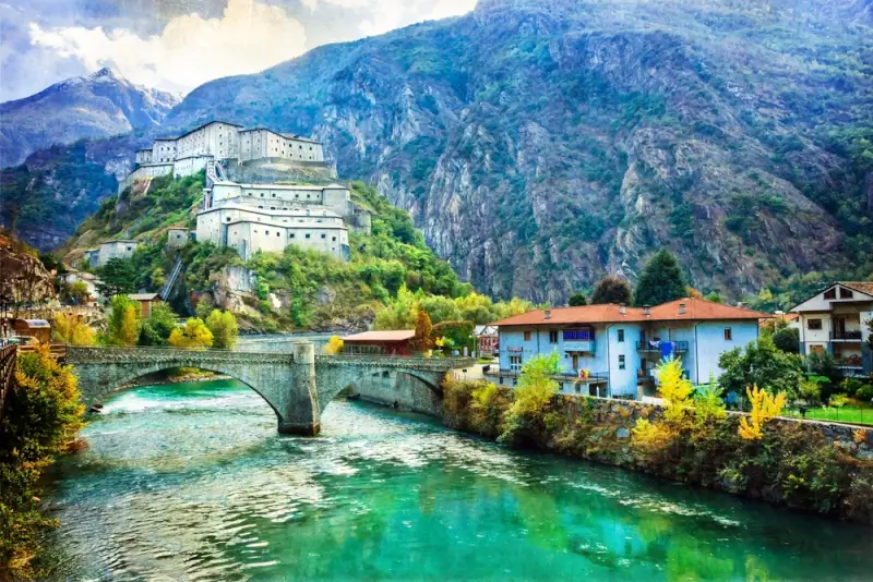 Historic fortress perched on hillside above stone bridge and turquoise river in Aosta Valley Italian Alpine village