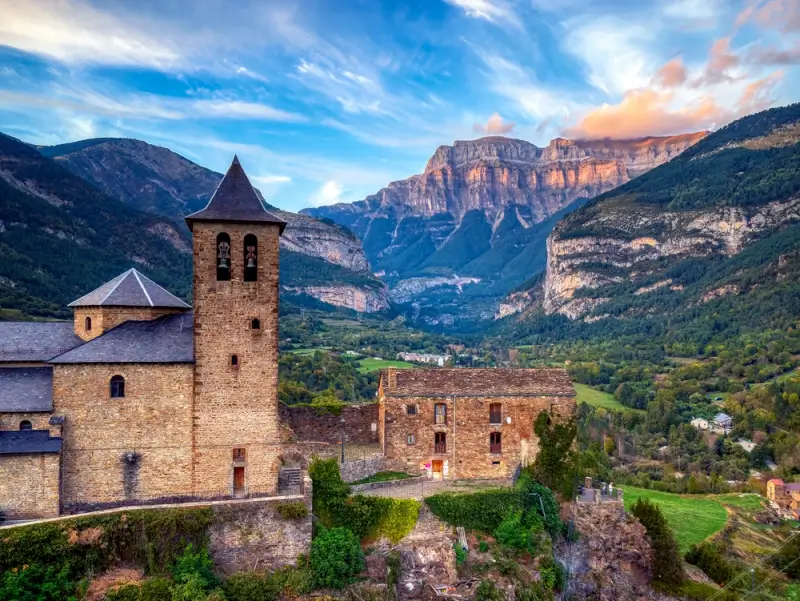 Historic stone church with bell tower overlooking mountain village at sunset with layered rock cliffs and green valley below