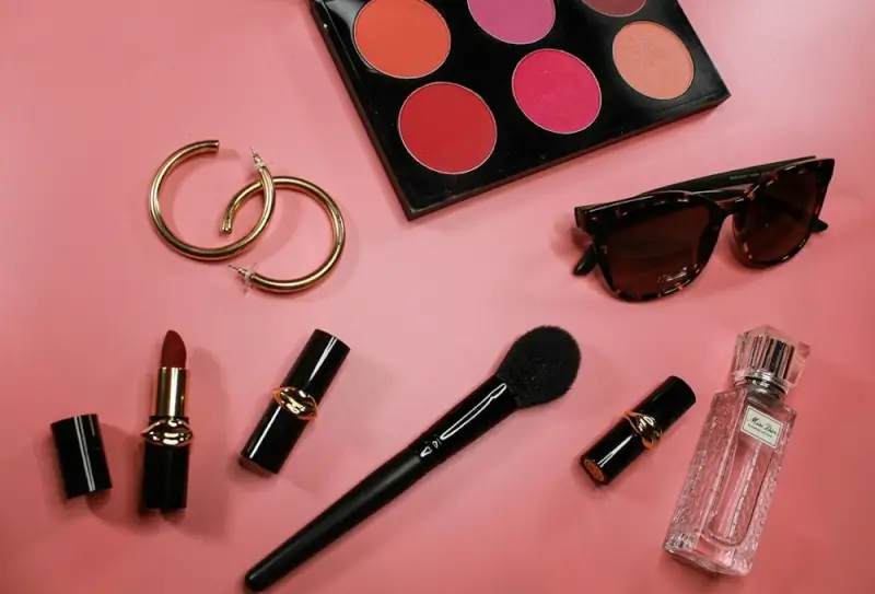 Beauty essentials flatlay with perfume bottle, makeup palette, lipsticks, and accessories on pink background