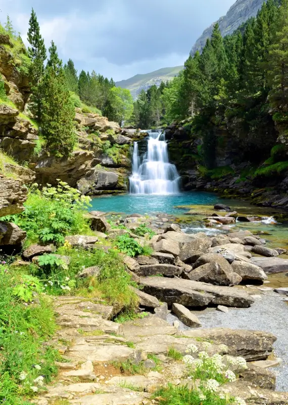 Cascading waterfall flowing into turquoise pool surrounded by moss-covered rocks, lush green forest, and mountain peaks in distance