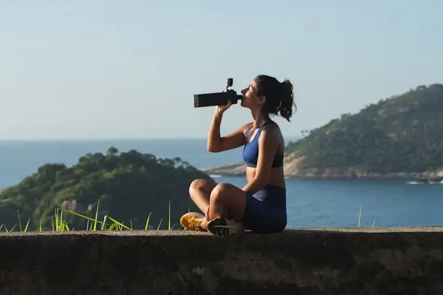 Woman in athletic wear drinking from black water bottle while sitting outdoors with ocean view