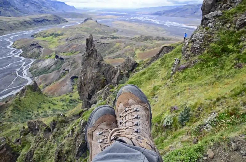 Hiking boots resting on moss-covered cliff edge overlooking Þórsmörk Valley with dramatic rock formations, glacial rivers, and a distant hiker in blue in Iceland