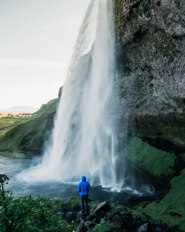 Solo hiker in blue jacket standing on moss-covered rocks beneath a towering waterfall cascading over volcanic cliffs in Iceland