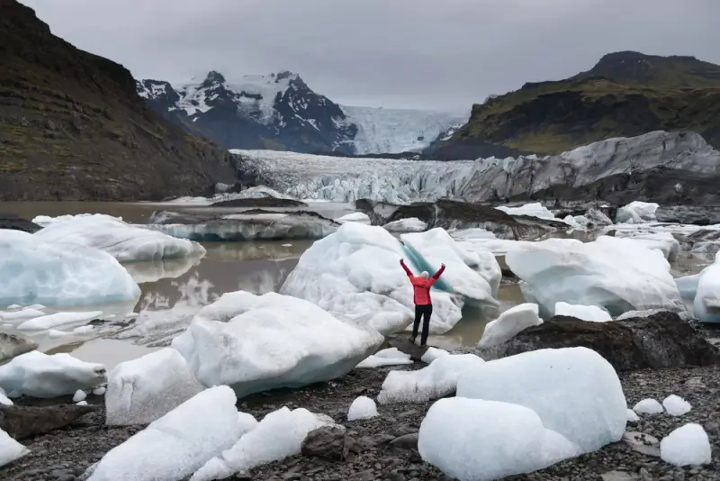 Woman traveler in red jacket with arms raised standing among white and blue icebergs at a glacial lagoon with glacier and mountains in the background in Iceland