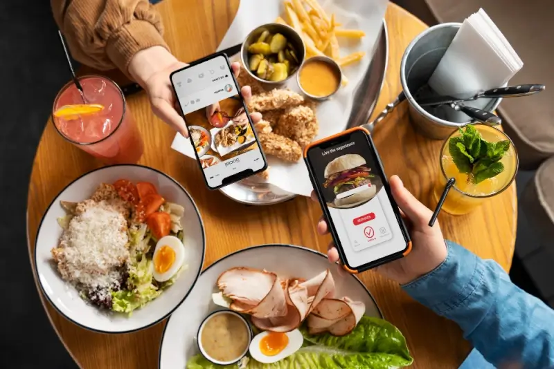 Two people sitting at a restaurant table using mobile food-ordering apps on their phones, surrounded by plates of salads, chicken tenders, fries, and colorful drinks.