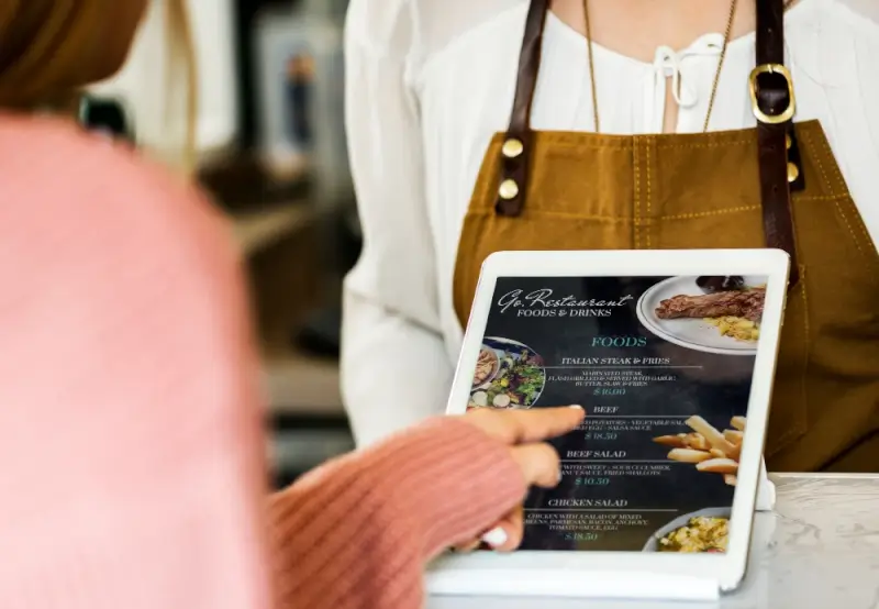 Customer pointing at a digital tablet menu held by a staff member in an apron, illustrating how a marketing plan for a restaurant can include modern digital ordering tools.