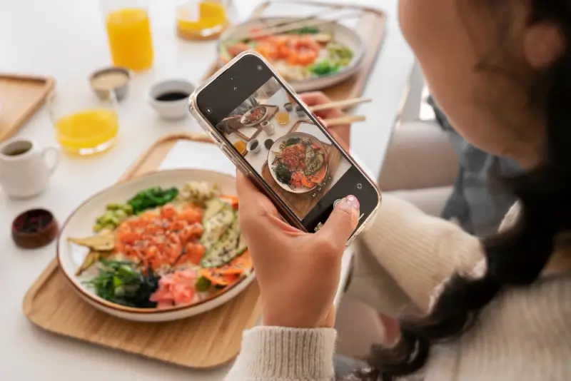 Person taking a photo of a colorful bowl of food with a smartphone, with plates of fresh ingredients and drinks arranged on a table