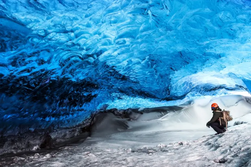 Hiker with orange helmet and backpack crouching at the entrance of a brilliant blue glacier ice cave in Iceland