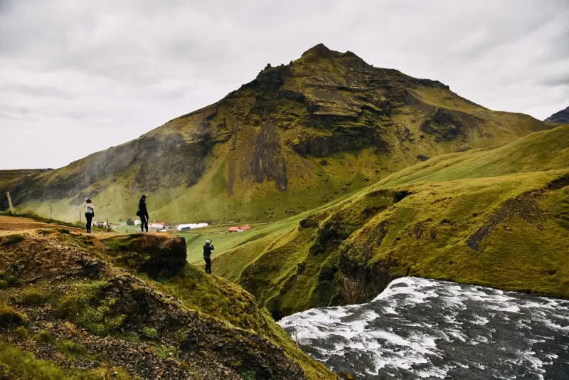 Iceland hiking trails along moss-covered cliffs near Skogafoss waterfall with hikers on the path and green volcanic mountains in the background