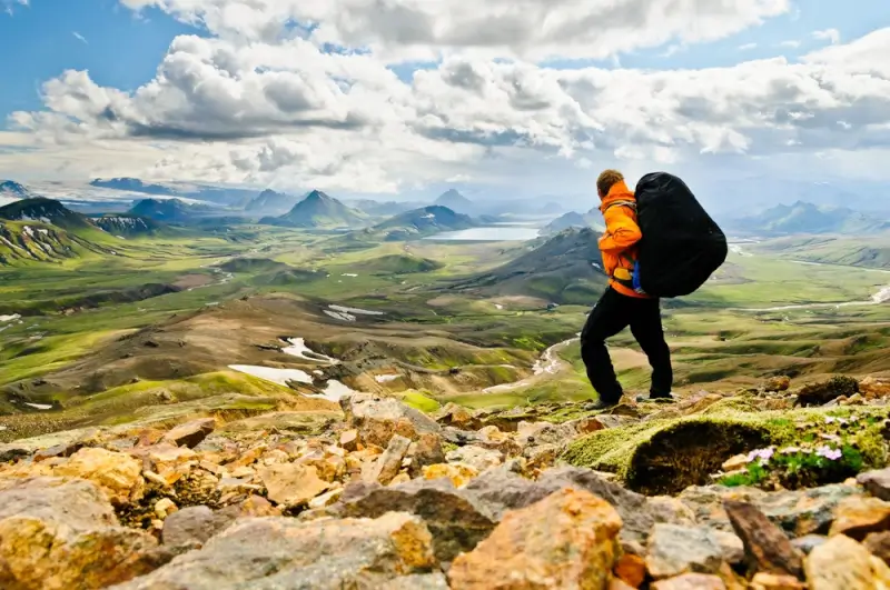 Hiker with orange jacket and large backpack walking on rocky trail overlooking vast volcanic valleys and mountains in Iceland's highlands