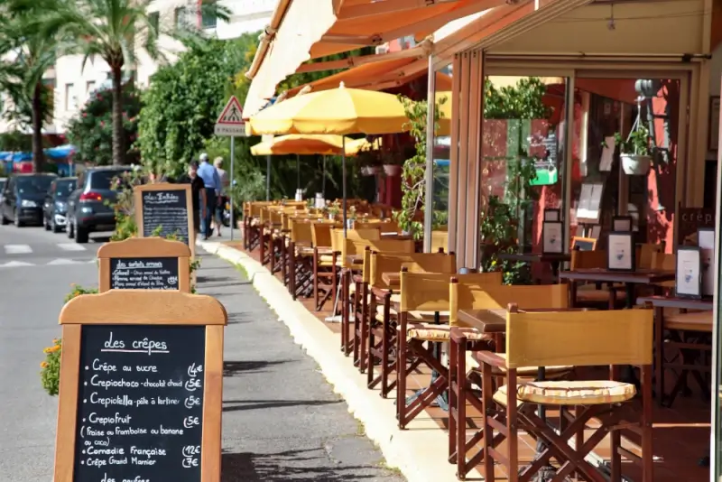 Outdoor café with rows of wooden tables and chairs under bright yellow awnings, featuring chalkboard menus on the sidewalk and a lively street scene in the background.