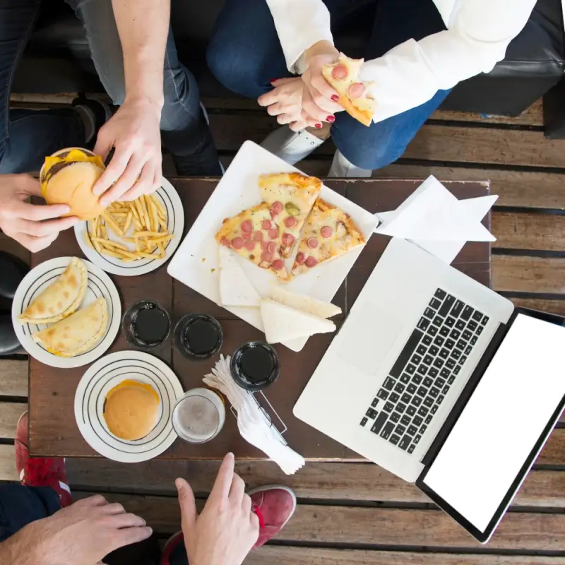 Top view of three people sharing fast food—including pizza slices, burgers, fries, and drinks—around a wooden table with an open laptop.