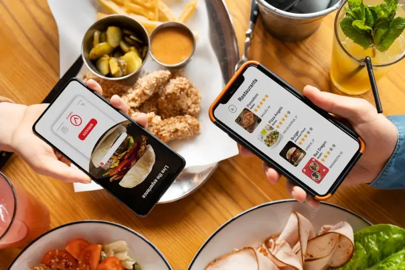 Two people holding smartphones showing food-delivery and restaurant review apps while eating fried chicken, pickles, salads, and drinks at a restaurant table.