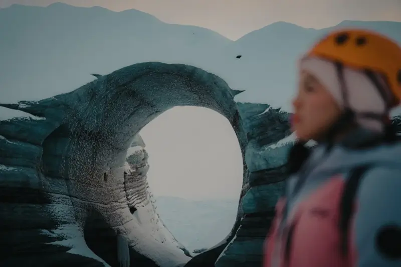 Solo traveler in orange helmet standing beside a circular ice formation with mountain views in winter Iceland